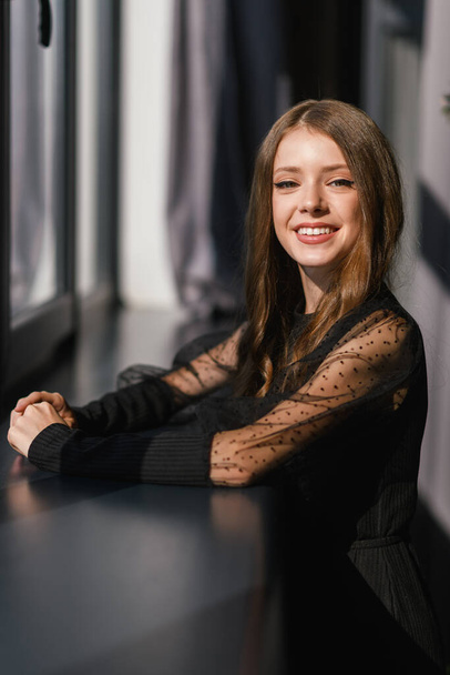Portrait of an attractive smiling teenage girl with her hands folded on the window sill. Girl in a black dress. - Photo, Image