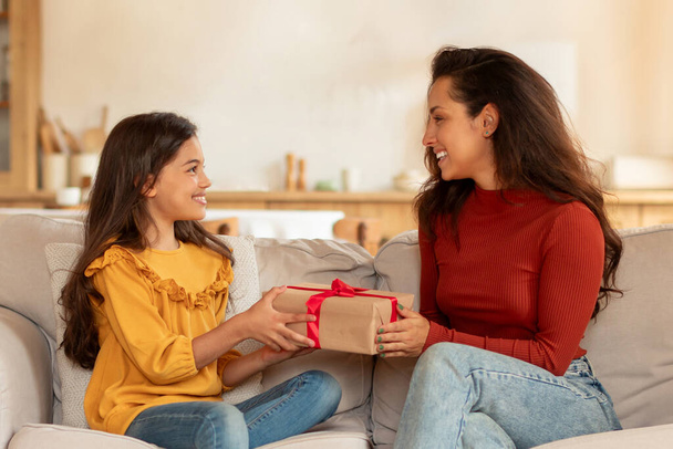 Prezent niespodzianka dla córki na urodziny. Radosna arabska mama daje zapakowane prezent pudełko dla nastolatki córka, siedzi na kanapie, gratulacje i świętowanie rodzinne wakacje w domu - Zdjęcie, obraz