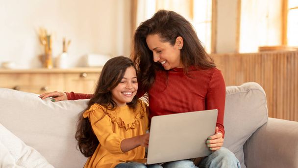 Mãe Oriente Médio alegre e filha estudante usando laptop na sala de estar moderna, sentado com computador, navegando na Internet e se divertindo on-line em casa no fim de semana. Panorama - Foto, Imagem