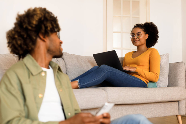Cónyuges afroamericanos felices navegando por la web a través de Smartphone y Laptop, pasando tiempo en línea en la sala de estar en casa. Hombre Mensajes de texto en el teléfono móvil, mientras que la esposa de trabajo en línea a través de la computadora, enfoque selectivo - Foto, Imagen