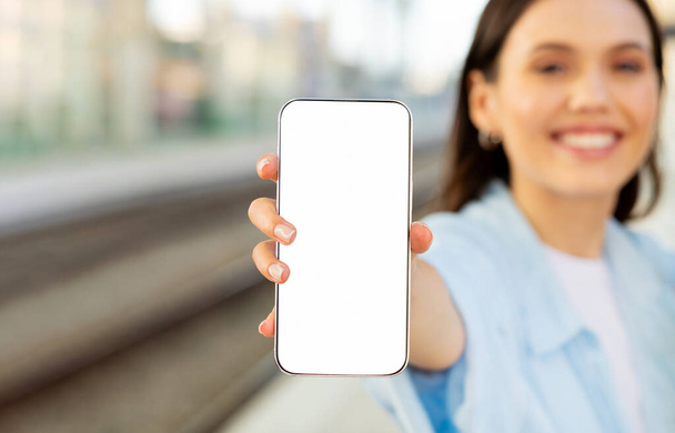 Ευτυχής νεαρή ευρωπαϊκή κυρία δείχνουν smartphone με άδεια οθόνη, στο σιδηροδρομικό σταθμό ή το αεροδρόμιο, με online εισιτήριο, κοντά. Συσκευή για το ταξίδι, χάρτης, συνιστά διαφήμιση και προσφορά, ιστοσελίδα, εφαρμογή σύσταση - Φωτογραφία, εικόνα