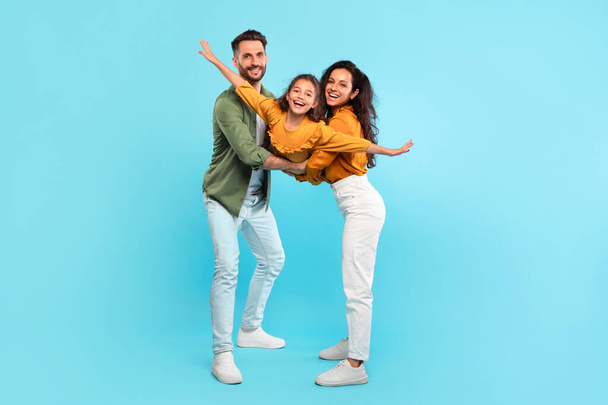 Excited parents playing with daughter holding her in arms posing on blue background, full length, family having fun together, girl spreading hands like plane - Photo, Image