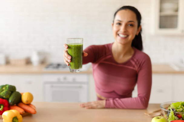 Zdravá detoxikační dieta. Fitness Lady nabízí Green Smoothie pití, držení skla a natahování ruku do kamery v moderní kuchyni doma, hubnutí a těší hubnutí recepty. Selektivní zaměření - Fotografie, Obrázek