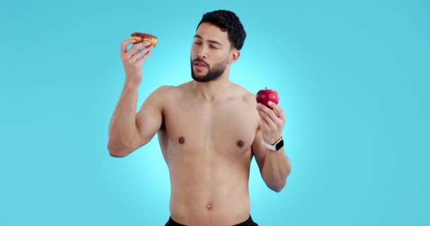 Voeding, man en shirtloos met appel en donut voor gezondheidszorgkeuzes of optie in studio op blauwe achtergrond. Fitness, portret en persoon met gezond eten, fruit of dieetadvies voor wellness. - Video