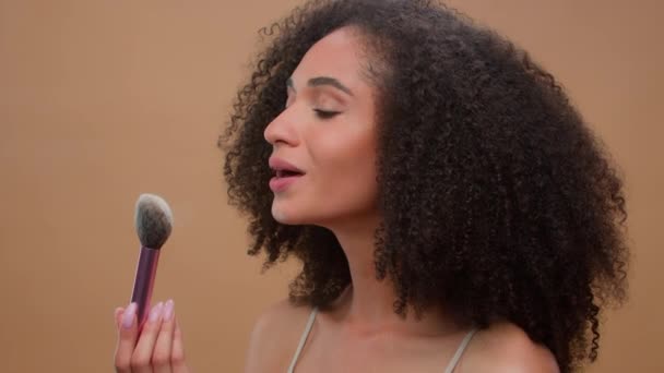 Close up portrét africký americký etnický žena model dívka drží štětec odfouknout prach aplikovat tón kůže prášek na nos usmívat se na kameru na béžové studio pozadí krásná dáma kosmetika make-up - Záběry, video