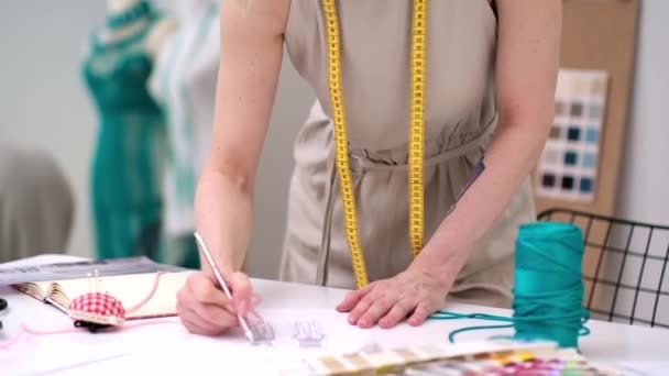デザイナーは,ファッションアトリエの熟練したドレスメーカーで働く紙に鉛筆で将来の女性のドレススケッチを描画し,ショップのクローズアップで職場で新しい衣服モデルを作成します - 映像、動画