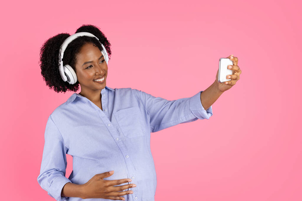 Тысячелетняя чернокожая дама с животом в беспроводных наушниках любит музыку, наслаждается свободным временем, делает селфи на телефоне, изолированном на розовом фоне, в студии. Стиль жизни, блог и социальные сети - Фото, изображение