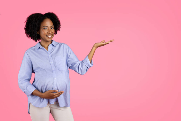 Θετική, χιλιετή μαύρη έγκυος γυναίκα που αγγίζει τη μεγάλη κοιλιά, κρατάει αντίγραφο χώρου, απολαμβάνει τη μητρότητα, απομονωμένη σε ροζ φόντο, στούντιο. Προτείνει για αναμένουν παιδί, πώληση, διαφήμιση, προσφορά - Φωτογραφία, εικόνα
