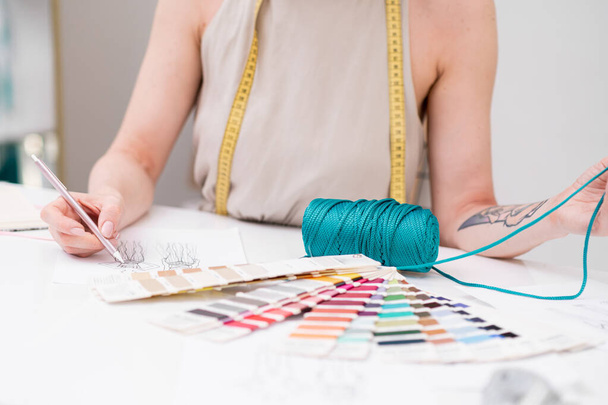 Dressmaker dessine des vêtements tenant le fil vert sur le lieu de travail dans l'atelier de mode créateur de vêtements crée un croquis de vêtement et choisit le matériau dans le studio de couture gros plan - Photo, image