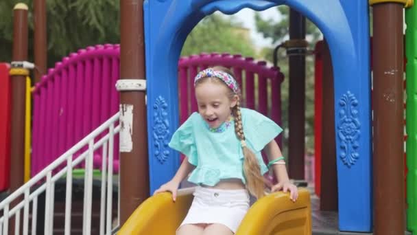 Sonriente niña paseos diapositiva de plástico en el laberinto de niños seguros parque infantil feliz niño con trenzas juega atracción en el parque público urbano colegiala disfruta deslizándose - Metraje, vídeo