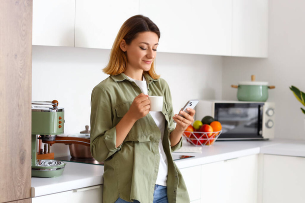Fiatal nő csésze forró kávé segítségével mobiltelefon a konyhában - Fotó, kép