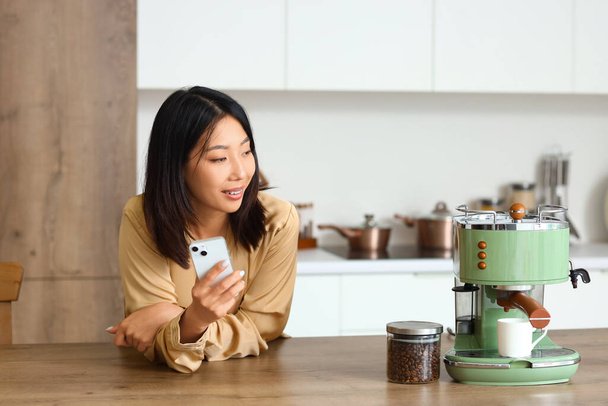 Hermosa mujer asiática con teléfono móvil y cafetera en la cocina - Foto, imagen