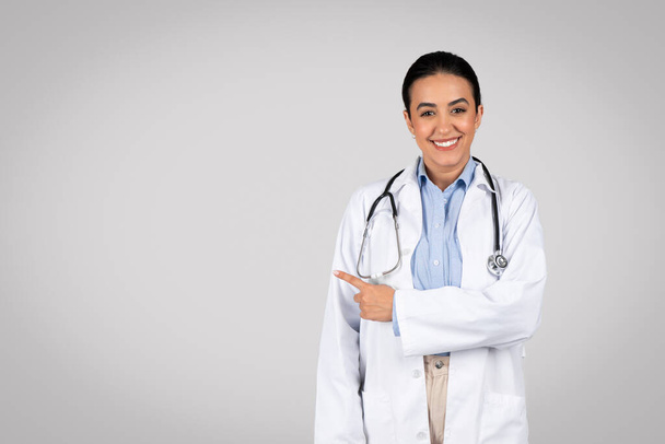 Informação útil. Retrato de alegre médico latino feminino apontando para longe no espaço de cópia em pé isolado sobre fundo de estúdio cinza, sorrindo para a câmera. Médico usando estetoscópio - Foto, Imagem