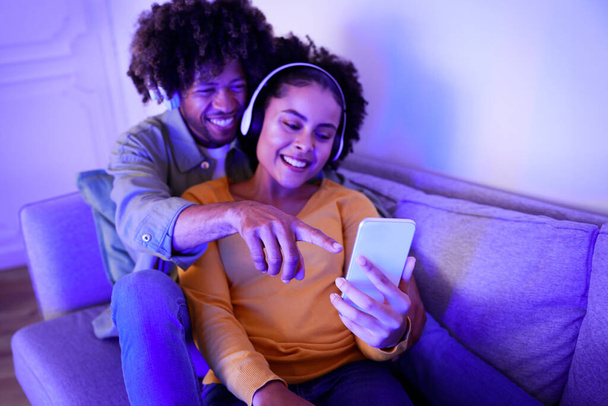 Χαμογελώντας African American Ζευγάρι Αγκαλιάζοντας και απολαμβάνοντας τη μουσική σε απευθείας σύνδεση στο έξυπνο τηλέφωνο, κάθισε στον καναπέ στο σπίτι, Lit με μπλε φως. Άντρας που δείχνει τις συζύγους Κινητό τηλέφωνο Περιήγηση στο Internet, επιλεκτική εστίαση - Φωτογραφία, εικόνα