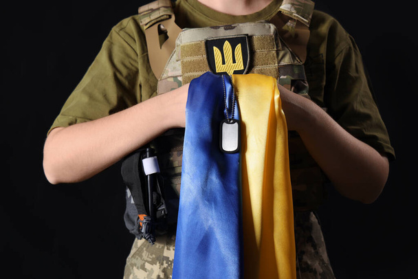 Joven soldado con bandera de Ucrania y etiqueta militar sobre fondo oscuro - Foto, Imagen