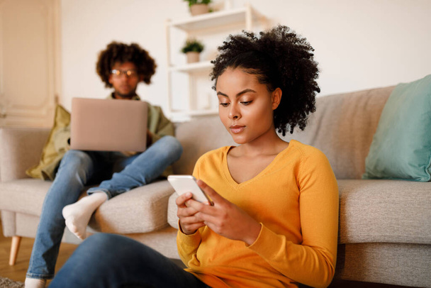 Familia conectada a Internet. Mujer afroamericana enfocada en mensajes de texto en el teléfono celular mientras el marido trabaja en el ordenador portátil en casa, joven pareja seria que participa en el ocio en línea. Tecnología. Enfoque selectivo - Foto, Imagen