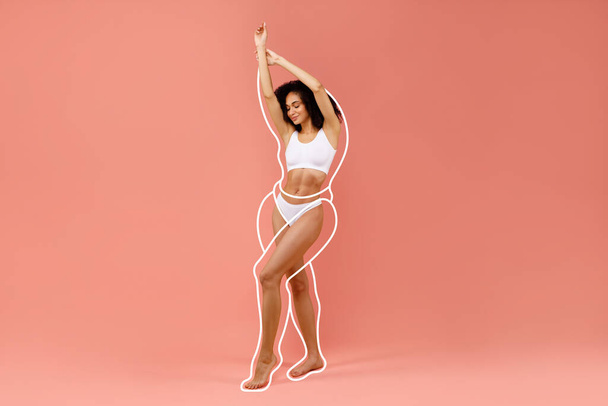 Heiße junge schwarze Frau posiert in weißer Unterwäsche, hebt die Hände, zeigt schöne Kurven ihres Körpers, steht auf rosa Studiohintergrund, zeigt Wiegeergebnisse, Kopierraum, volle Länge - Foto, Bild
