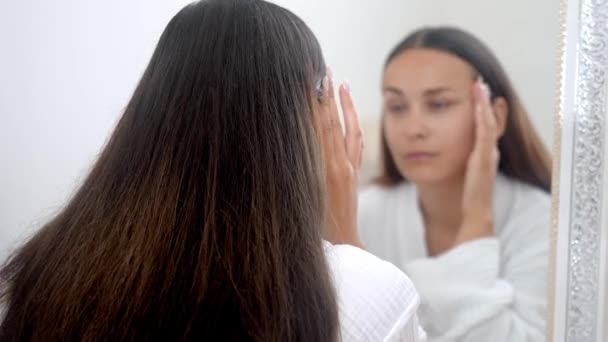 Eine Frau, zu Hause im Bademantel, vertieft sich in ein Schönheitspflegekonzept und untersucht sich selbst im Badezimmerspiegel.  - Filmmaterial, Video