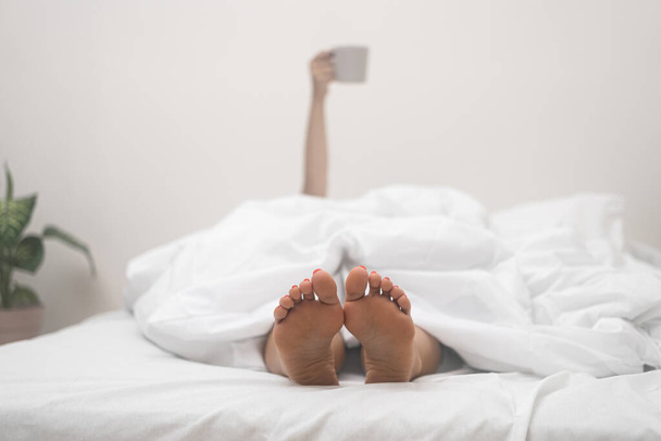 Mujer con los pies desnudos descansa en la cama ligera con taza de café en la mano que sufre de insomnio enfermedad incapacidad para dormir en condiciones cómodas en el hogar - Foto, Imagen