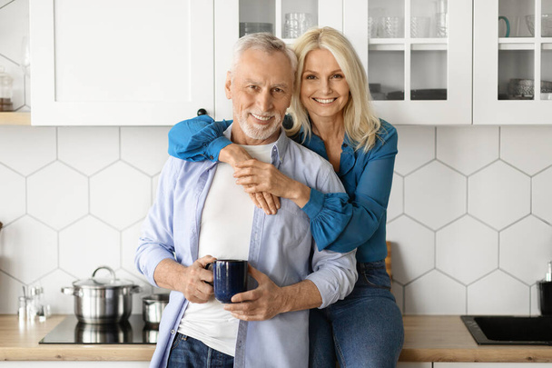 Porträt eines romantischen attraktiven Seniorehepaares, das in der gemütlichen Küche steht, schöne reife Ehepartner, die zusammen frischen Morgenkaffee trinken, sich umarmen und in die Kamera lächeln, Kopierraum - Foto, Bild