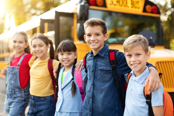 Happy Multiethnic dzieci pozowanie na zewnątrz w pobliżu autobusu szkolnego, Wesoły nastolatek chłopców i dziewcząt z plecakami stojących na zewnątrz, Obejmuje i uśmiecha się do kamery, Korzystanie z nauki razem, zbliżenie - Zdjęcie, obraz