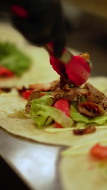 A chef in a restaurant prepares a shawarma or doner kebab, putting greens and pieces of juicy vegetables and hot peppers with red tomatoes and chicken on a thin pita spread on the table - Footage, Video