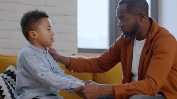 Őszinte szülői szeretet. Gondoskodó afro-amerikai apa beszél a feldúlt kisfiával, elmagyarázza neki az élet pillanatait és megnyugtatja őt, otthon ül együtt, oldalnézetből - Felvétel, videó