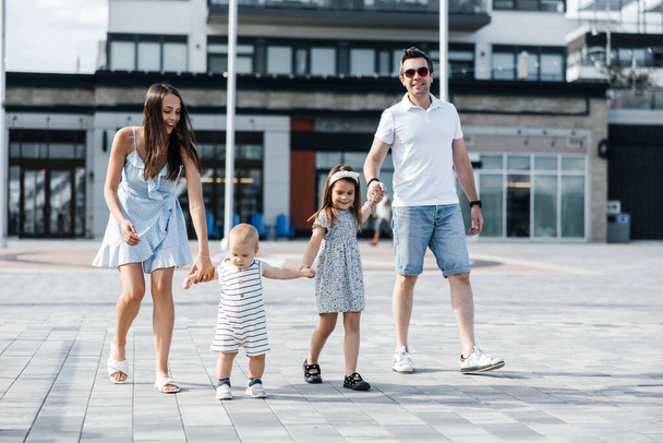 A happy young family walks on the street holding hands. Mom dad their daughter and son are holding hands and smiling. A cheerful walk in the fresh air of a family of four in summer clothes - Photo, Image