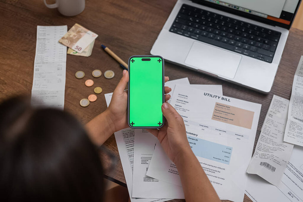 E-Paymen: A womans hand, securing a mobile phone with a chroma key screen, rules the wooden desk surrounded by utility bills, money, and an online banking app.  - Photo, Image