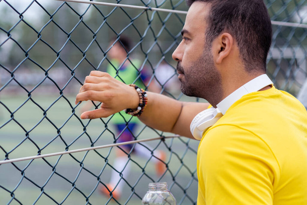 Любитель спорта с наушниками наблюдает за тренировками местной футбольной команды на поле через металлическую решетку забора, заинтересованный мужчина смотрит на футболистов на городской арене - Фото, изображение