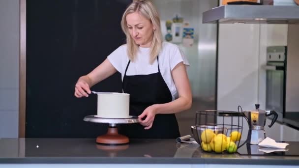 Šťastná žena se těší práci na domácí vlastní dort pro svatební oslavu profesionální cukrář pokrývající pečivo s bílou tunikou stojící na kuchyňském stole - Záběry, video