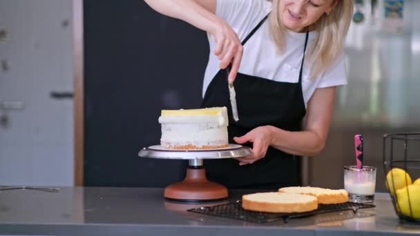 Tyytyväinen makeiskauppias hymyilee samalla, kun levitetään kakkukerroksia tuoreen orgaanisen kerman kanssa blondi nainen mustassa esiliinassa seisomassa keittiön pöydässä nauttien leivonnaisista asiakkaille - Materiaali, video