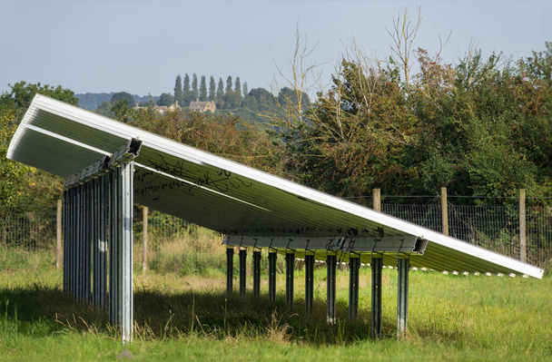 Vista lateral y detallada de paneles solares, unidades de trabajo, que brillan y reflejan la luz del sol al final de la tarde, mostrando casas rurales distantes en el horizonte, hierba verde de verano . - Foto, Imagen