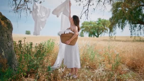 Country lady taking dry linen from rope at summer field. Young brunette put clean washed laundry to wicker basket at countryside nature alone. Tender calm woman in courtyard. Village lifestyle concept - Footage, Video