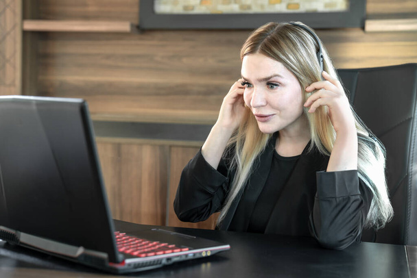 Επιχειρηματίας γυναίκα σε μαύρο κοστούμι με ακουστικά κάθεται μπροστά από το laptop στο γραφείο - Φωτογραφία, εικόνα