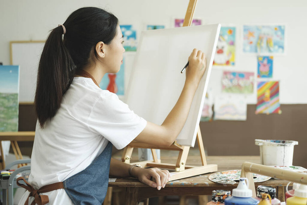 Fiatal női művész rajzolja vagy festi a rajzát vászonra egy műhelyben. Egy tinédzser lány, aki szereti a művészetet és a rajzot, időt vesz igénybe, hogy nagy szándékkal alkossa meg az akvarelljeit vásznon. - Fotó, kép