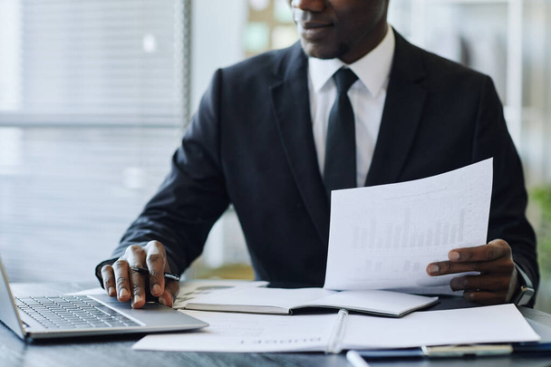 Обрізаний знімок молодого чорного бізнесмена в костюмі, використовуючи ноутбук на робочому місці в офісі і тримаючи контракт, простір для копіювання - Фото, зображення