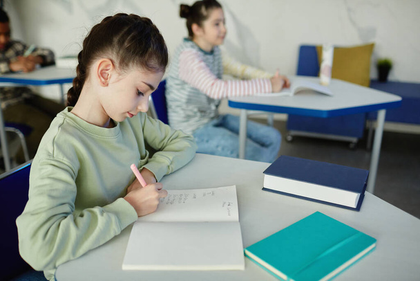 Ritratto laterale di giovane studentessa che scrive nel quaderno mentre è seduta alla scrivania durante la lezione, copia lo spazio - Foto, immagini