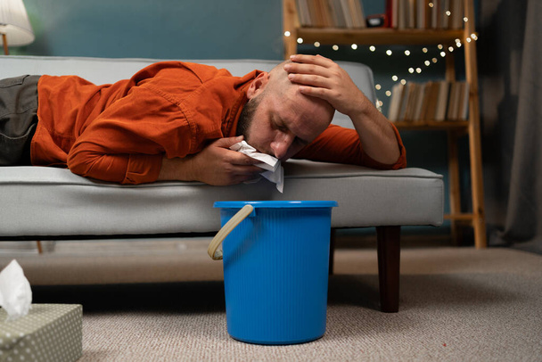 Un homme vomissant dans un seau sur le canapé à la maison. Intoxication, empoisonnement et nausées concept. Gros plan - Photo, image