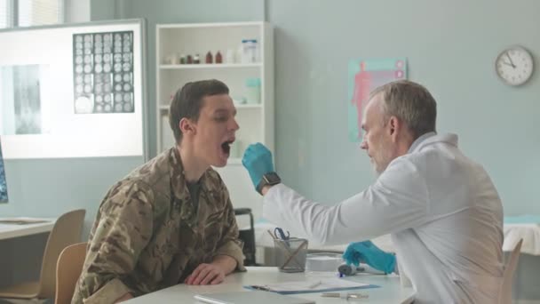 Média foto de médico militar de meia idade vestindo jaleco branco e luvas descartáveis fazendo teste de cotonete oral para soldado durante consulta médica - Filmagem, Vídeo