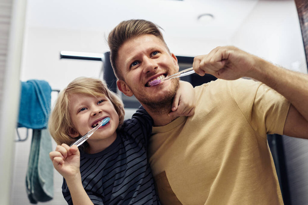 Низкоугольный портрет игривого отца и сына, чистящих зубы вместе утром и смотрящих в зеркало - Фото, изображение