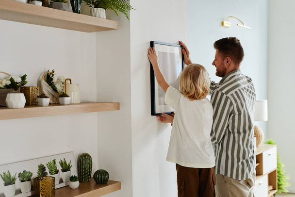 Back view portrait of father and son hanging picture together in minimal home interior decorated with succulents, copy space - Photo, Image