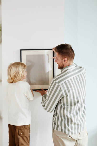 Vertical back view portrait of father and son hanging picture together and looking at each other - Photo, Image