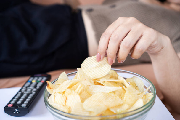 kobieta leżąca na kanapie jedząca chipsy ziemniaczane, niezdrowy tryb życia  - Zdjęcie, obraz