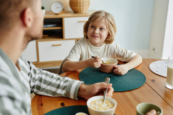 Alto ángulo retrato de padre e hijo disfrutando del desayuno juntos en la mañana con enfoque en lindo chico rubio sonriendo - Foto, Imagen