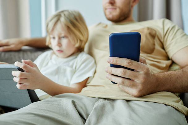 Primer plano del padre sosteniendo el teléfono inteligente con el niño pequeño usando la tableta en el fondo, espacio de copia - Foto, imagen