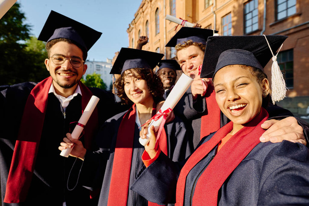 Heureux diplômés faisant portrait selfie en robes contre l'université en plein air - Photo, image