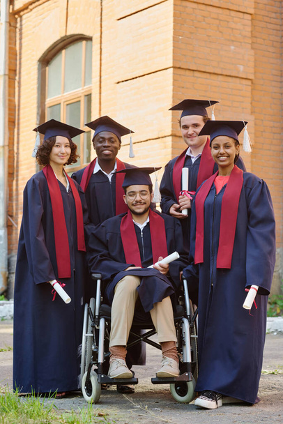 Immagine verticale di studenti laureati in vesti sorridenti davanti alla macchina fotografica mentre stanno all'aperto contro l'università - Foto, immagini