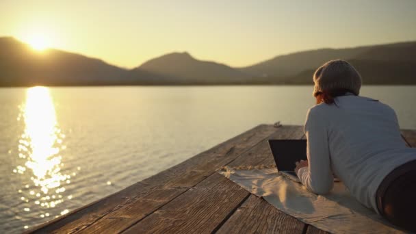 Ελεύθερη επιλογή εργασίας έννοια: μια νεαρή γυναίκα εργάζεται στον φορητό υπολογιστή της που βρίσκεται σε μια λίμνη ή αποβάθρα στη θάλασσα το ηλιοβασίλεμα σε ένα όμορφο φυσικό περιβάλλον. Βελτίωση της ποιότητας ζωής με το να ζει κανείς στη φύση - Πλάνα, βίντεο