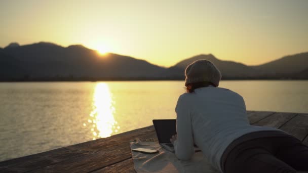 Freelance werkkeuze concept: een jonge boekenschrijfster schrijft op haar laptop terwijl ze bij zonsondergang op het dok van een meer of zee ligt, in een prachtige natuurlijke omgeving. Werken op afstand om beter te leven - Video